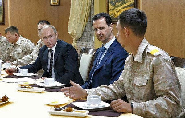 A handout photo released on the official Facebook page of the Syrian Presidency on December 11th shows Russian President Vladimir Putin and Syrian President Bashar al-Assad meeting with Russian military officers in the Russian air base in Hmeimim in Syria's Latakia province. [Syrian Presidency Facebook page/AFP]