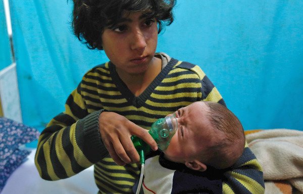 A Syrian boy January 22nd, 2018, in Douma holds an oxygen mask over the face of an infant following a reported gas attack. Since the beginning of the war in Syria in 2011, the government of Bashar al-Assad has been repeatedly accused by UN investigators of using chlorine gas or sarin gas against civilians. [Hasan Mohamed/AFP]