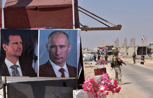 Members of Russian and Syrian forces stand guard near posters of Syrian President Bashar al-Assad and his Russian counterpart Vladimir Putin at the Abu Duhur crossing on the eastern edge of Idlib province on August 20th, 2018. [George Ourfalian/AFP]