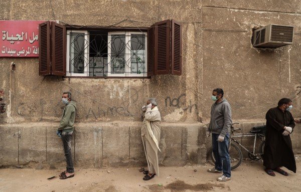 Egyptian men wearing masks wait outside a centre of NGO Egyptian Food Bank in Cairo to receive cartons of foodstuffs on April 5th, as the charity distributes aid to people who lost their jobs due to the coronavirus pandemic crisis. [Mohammed al-Shahed/AFP]