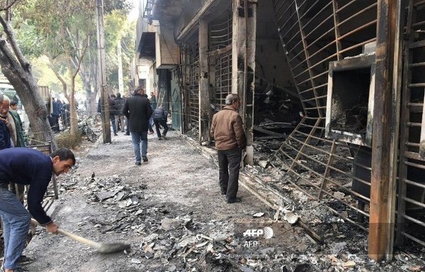 چند تن از ایرانیان در تاریخ ۱۷ نوامبر ۲۰۱۹ در اصفهان از مقابل یک بانک، که در جریان اعتراضات مردمی نسبت به افزایش قیمت بنزین به آتش کشیده و سوخته شده است، رد می‌شوند. [خبرگزاری فرانسه]