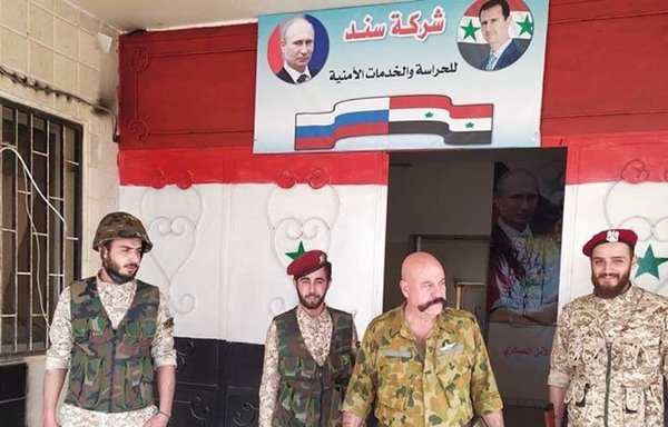 Brig. Gen. Raslan Esbar (2nd from right) stands with Sanad company personnel at a headquarters building beneath Russian and Syrian flags and portraits of the Syrian and Russian presidents. [Sanad]