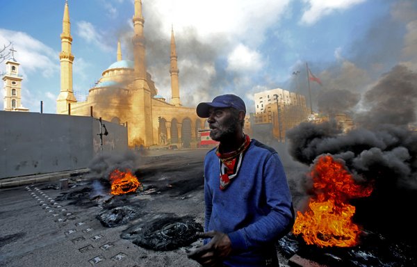 A man stands next to flaming tires at a make-shift roadblock set-up by anti-government demonstrators next to the Mohammed al-Amin Mosque in the Martyrs' Square in the centre of Lebanon's capital Beirut on March 8, 2021, during a protest against the deteriorating value of the local currency and dire economic and social conditions. [ANWAR AMRO / AFP]