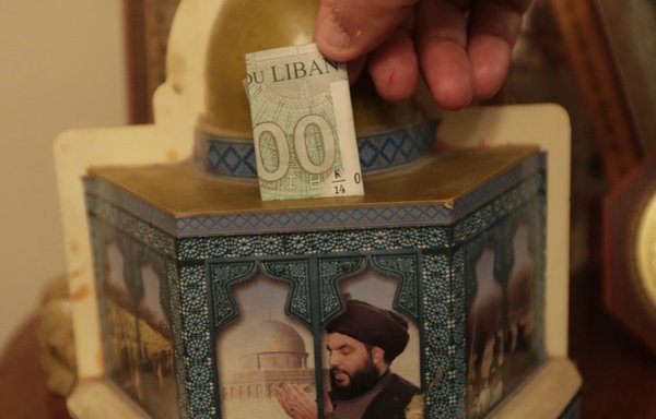 A man drops a banknote in a donation box bearing a portrait of Hizbullah chief Hassan Nasrallah in a southern suburb of Beirut on June 3, 2019. The militant group is coming under increasing financial pressure as a result of the sanctions imposed on it and on its sponsor Iran by the United States and its allies. [Anwar Amro/AA/AFP]