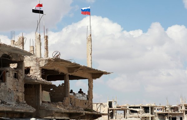 A Syrian and a Russian flag fly above a damaged building in Daraa, Syria on September 12, 2021. [Louai Beshara/AFP]